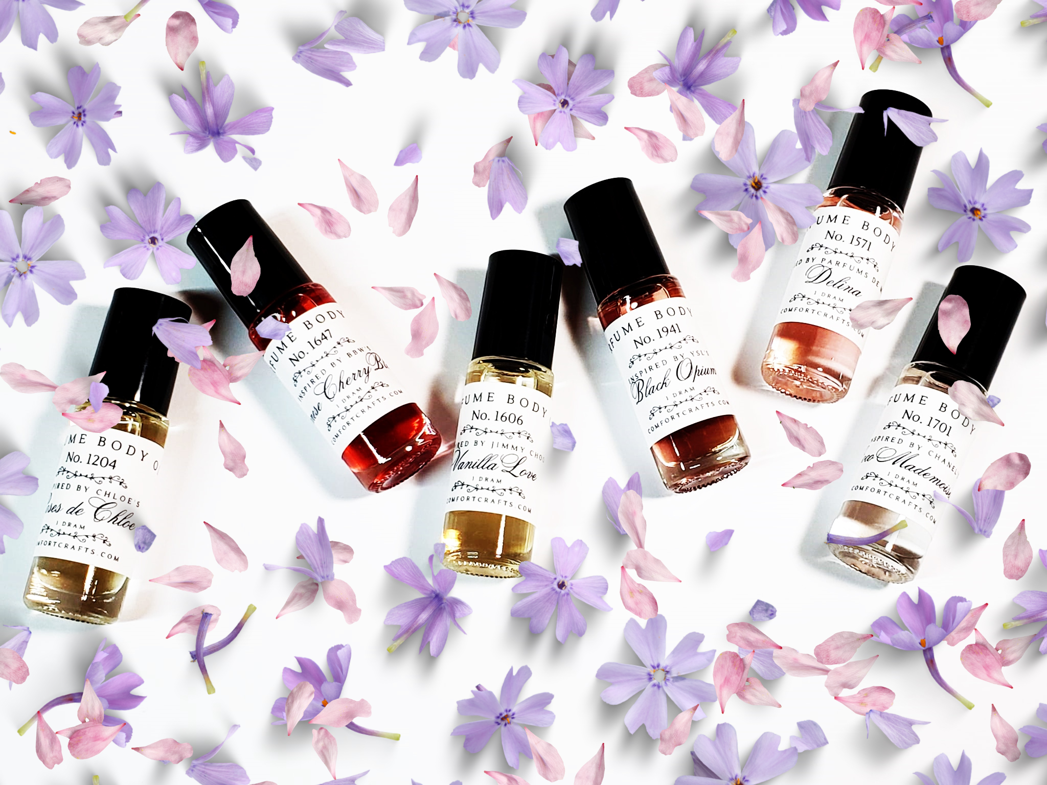 Six colorful dram size women's perfume oil bottles laying flat with pink and lavender colored flower petals around them.