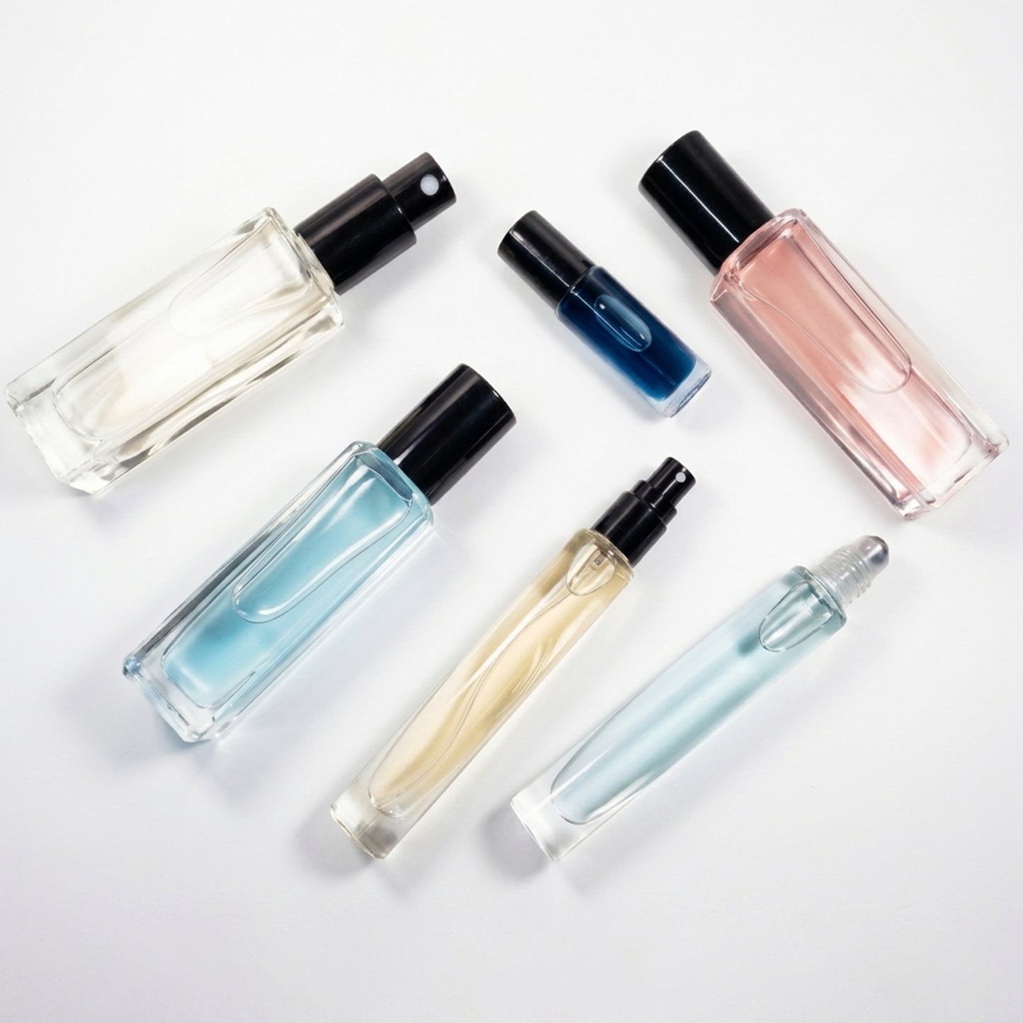 Six clear glass fragrance bottles and roller bottles with black and silver caps, filled with liquids in shades of pink, blue, yellow, and clear, arranged on a white background.