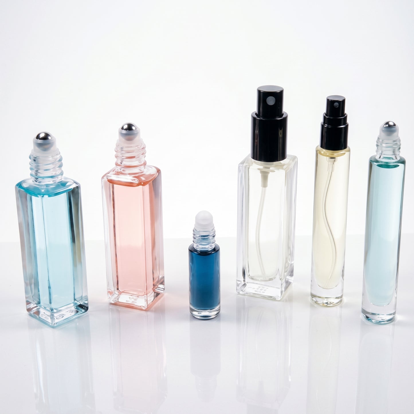 Six clear glass perfume or fragrance bottles of different shapes and sizes, some with black spray tops and one with a silver rollerball cap, filled with blue, pink, and pale liquid, arranged on a white reflective surface against a white background.