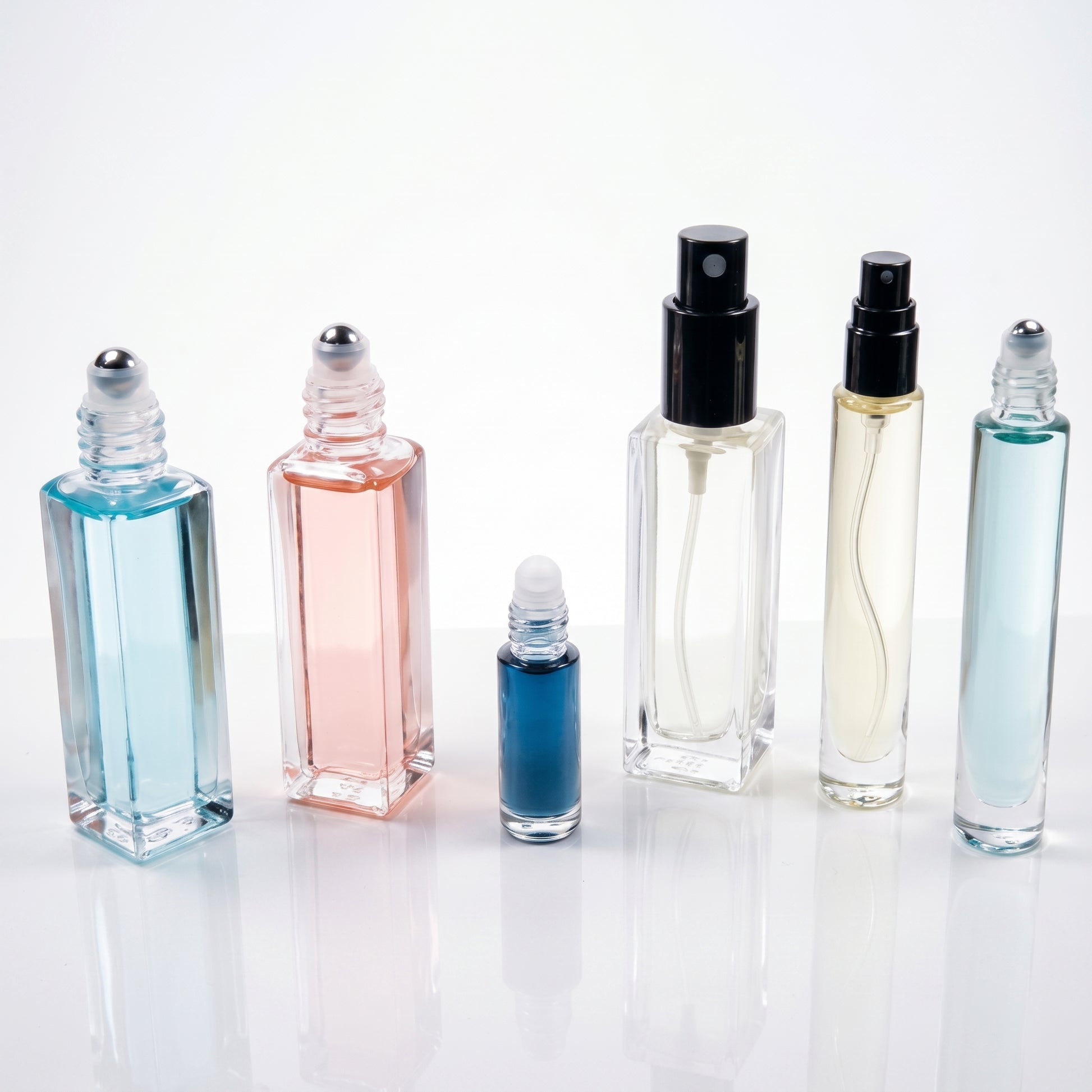 Six clear glass perfume or fragrance bottles of different shapes and sizes, some with black spray tops and one with a silver rollerball cap, filled with blue, pink, and pale liquid, arranged on a white reflective surface against a white background.